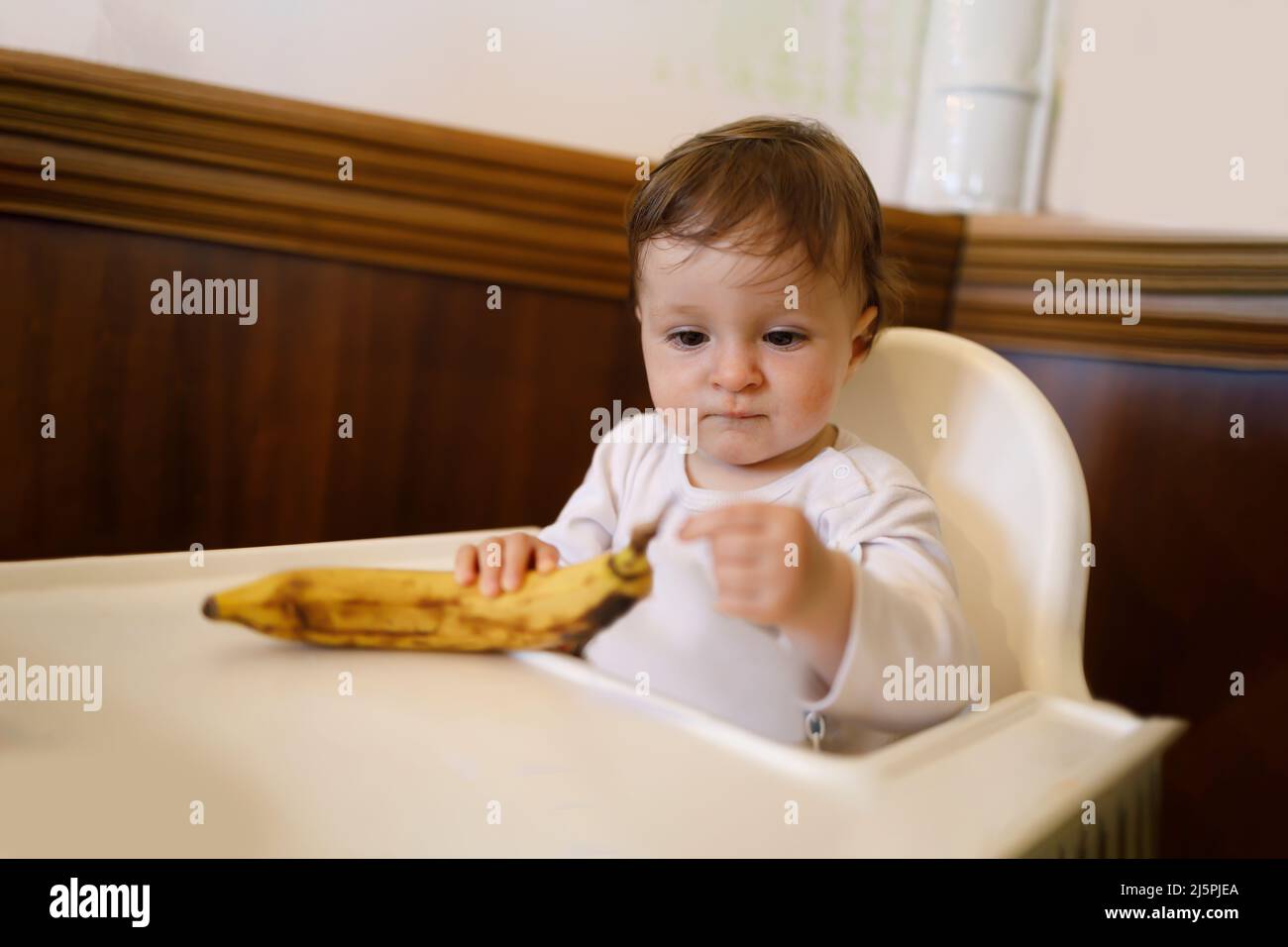 Kleines Kind, das am Esstisch sitzt und mit einer Banane spielt Stockfoto
