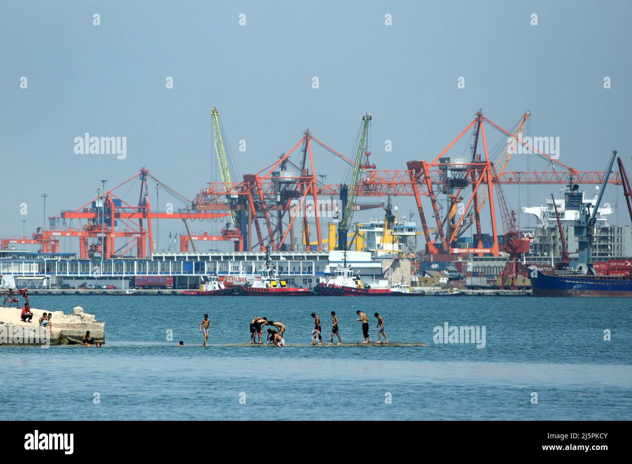 Türkische Jungen spielen an einem heißen Sommertag im türkischen Mersin auf einer Betonplattform im Mittelmeer. Im Hintergrund ist der Hafen von Mersin. Stockfoto
