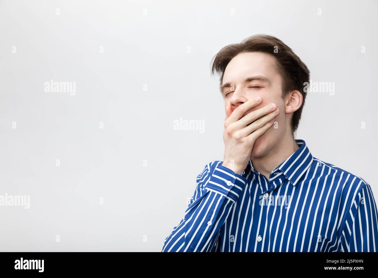 Junger Mann mit blau-weißem gestreiftem Knopfhemd, das den offenen Mund mit der Hand bedeckt, während er gähnte. Studio auf grauem Hintergrund aufgenommen Stockfoto