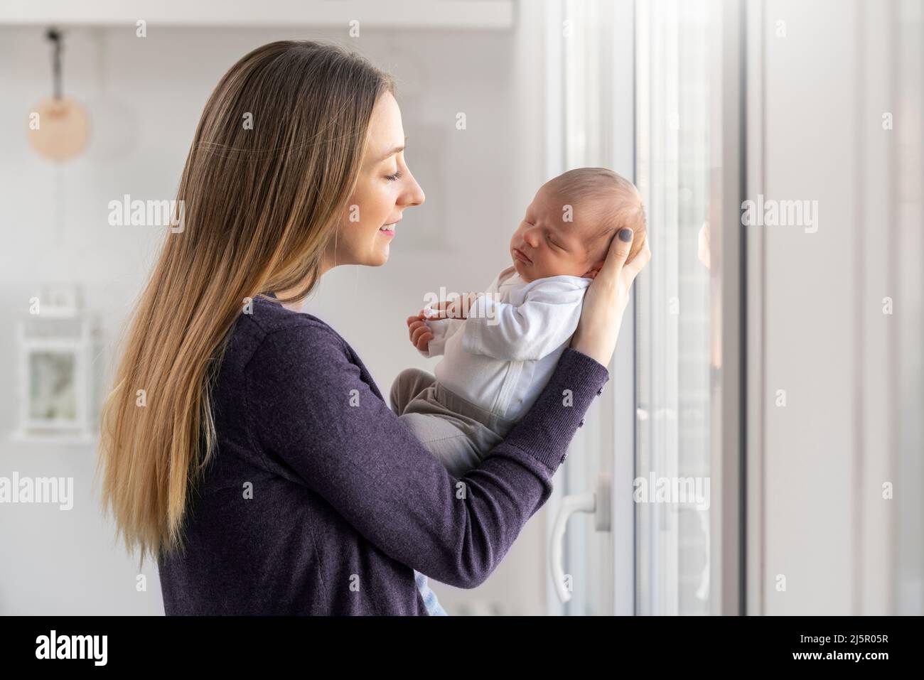 Junge Mutter mit ihrem neugeborenen Baby in den Armen zu Hause in der Nähe des Fensters Stockfoto