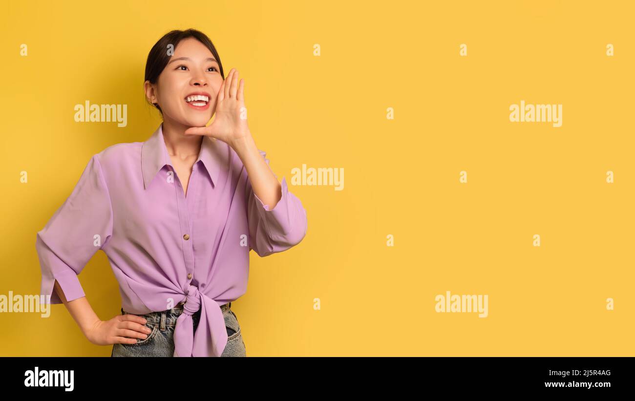 Junge asiatische Frau, die laut auf den Kopierraum schreit und die Hand am Mund auf gelbem Studiohintergrund hält, Panorama Stockfoto