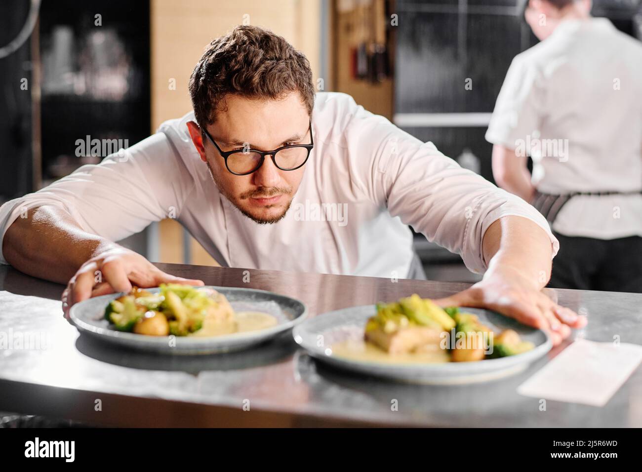 Ernsthafter Koch in Brillen, der Teller mit Hauptgericht auf der Theke ansah und sie für die Lieferung vorbereitete Stockfoto