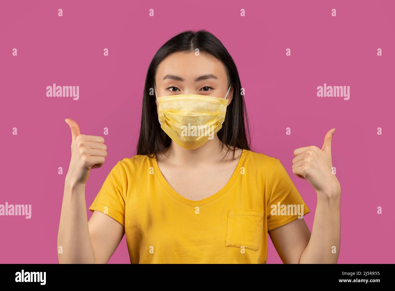 Porträt einer jungen glücklichen asiatischen Dame, die eine medizinische Gesichtsmaske trägt und die Daumen nach oben zeigt und auf violettem Hintergrund posiert Stockfoto