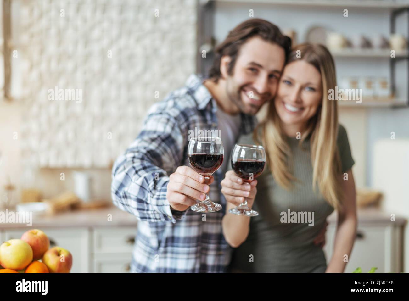 Frohes, tausendjähriges europäisches Paar umarmt sich, klinkende Gläser Wein und schaut euch die Kamera im skandinavischen Kücheninterieur an Stockfoto
