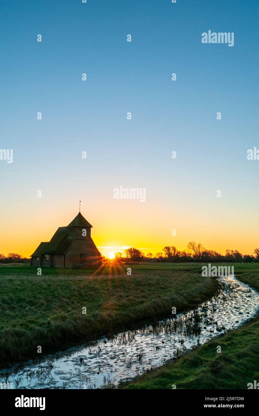 Die Kirche St. Thomas a Becket aus dem 13.. Jahrhundert am Romney Marsh, mit dem Sonnenaufgang in einem klaren Himmel, und im Vordergrund ein kleiner Bach mit Schilf. Winter. Stockfoto