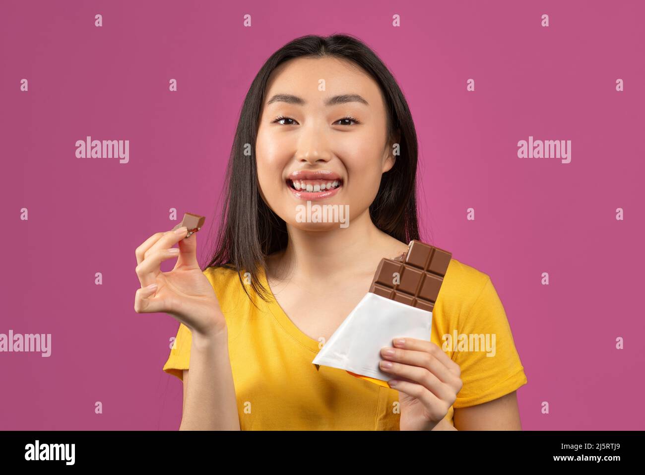 Fröhliche koreanische Dame, die Schokolade isst, leckeres Dessert genießt und die Kamera lächelt und über violettem Studiohintergrund steht Stockfoto