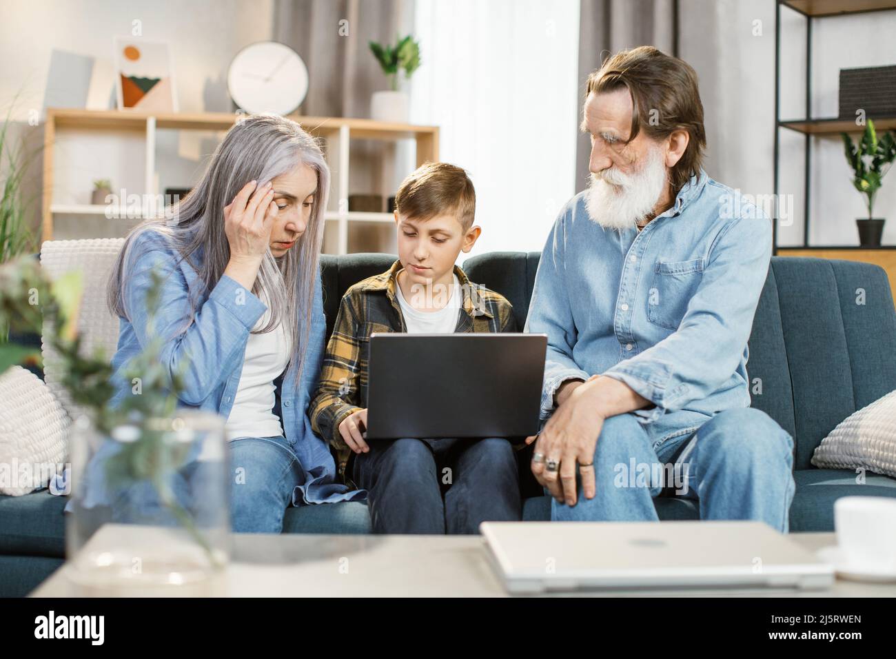 Enkel lehren ältere Menschen, mit Computer, Technologie und modernem Lebensstil im Internet zu surfen. Glücklicher kaukasischer Großeltern mit Enkelkind, das zu Hause auf dem Sofa sitzt und Laptop spielt Stockfoto