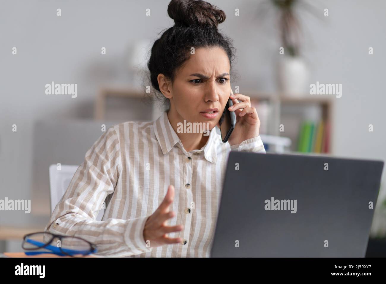 Verwirrte junge Geschäftsfrau, die mit einem Laptop über das Mobiltelefon spricht, Probleme mit dem Computer hat und im Büro arbeitet Stockfoto