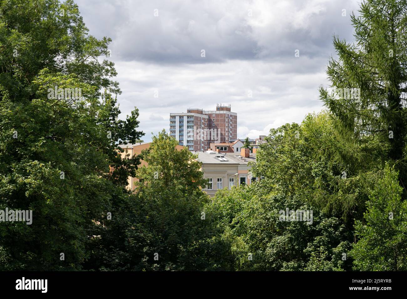 Altes Hochhaus in der Stadt vor kleinen Häusern. Großer dunkler Wolkenkratzer mit grünen Bäumen davor. Ostdeutsche Architektur Stockfoto