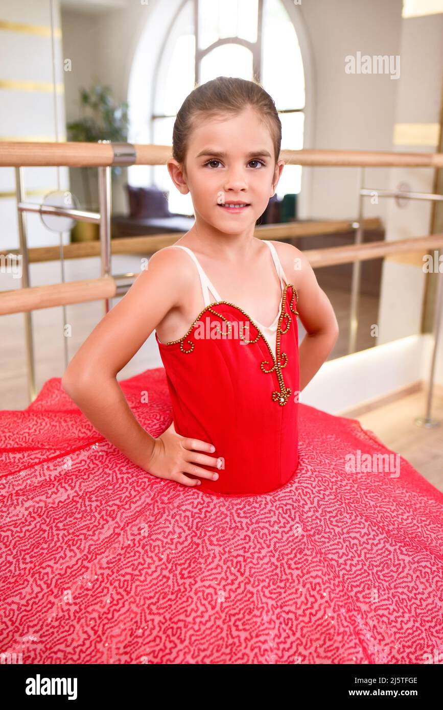 Niedliches Porträt einer schönen kleinen Ballerina in einem Performance-roten Kleid mit rosa Tutu. Sie lächelt, weil sie glücklich ist, ein Profi-Ball zu werden Stockfoto