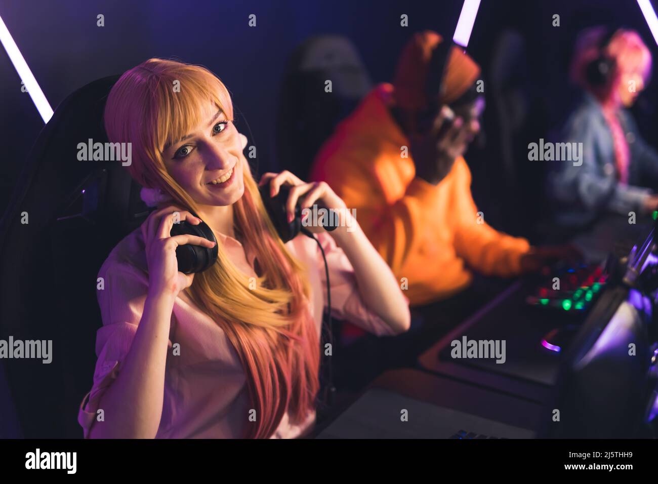 Bezauberndes kaukasisches langhaariges blondes Gamermädchen, das die Kamera anschaut, lächelt und ihr professionelles Headset aufsetzt. PC-Bang-Umgebung. Hochwertige Fotos Stockfoto