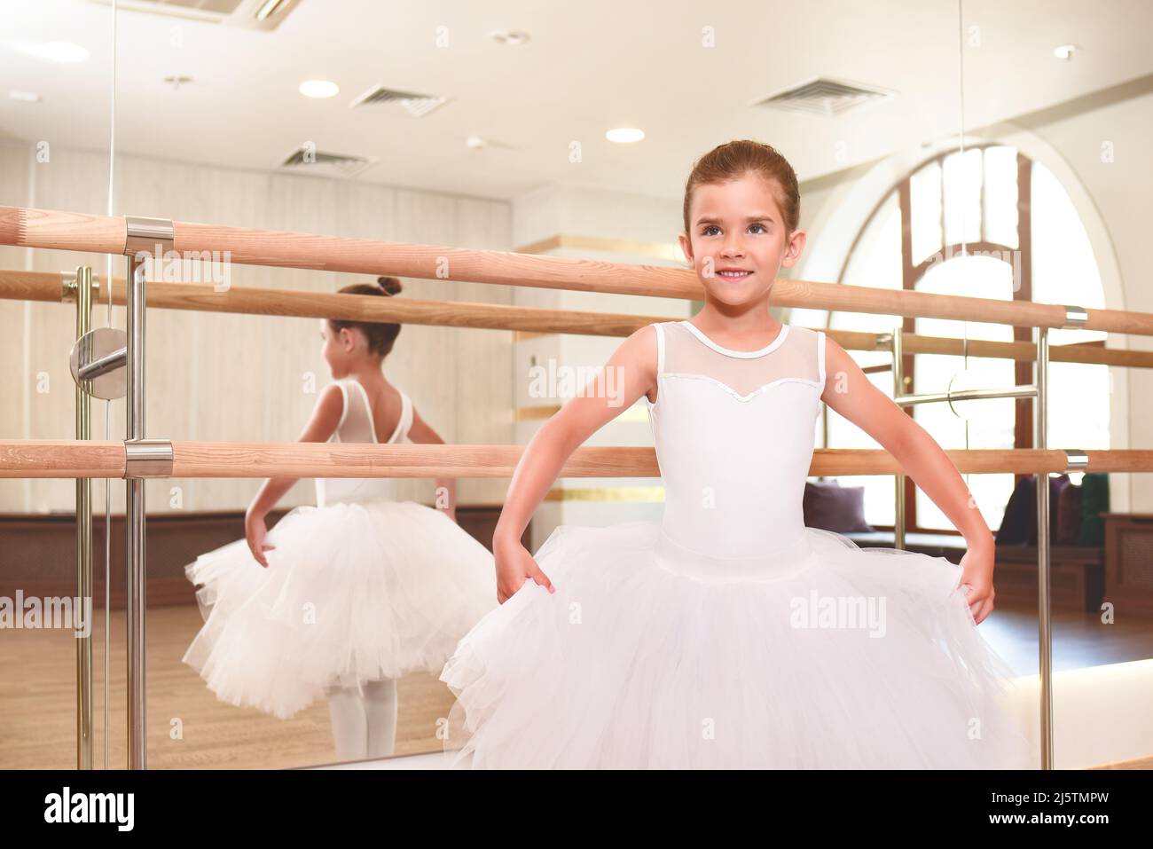 Eine niedliche kleine Ballerina in einem Tanzsaal mit großen Spiegeln hält begeistert die Ränder ihres weißen Tutus, sie ist bereit zu tanzen. Stockfoto