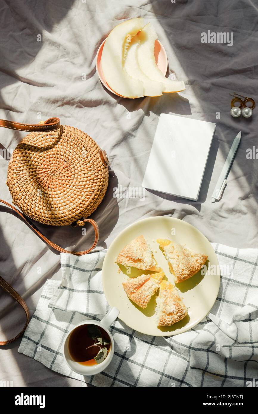 Frühstück im Bett auf Decke mit Tee, Obst und Kuchen. Wickertasche und Notizbuch. Hellgrüne Farbe. Inspiration für die Sommerstimmung. Stockfoto