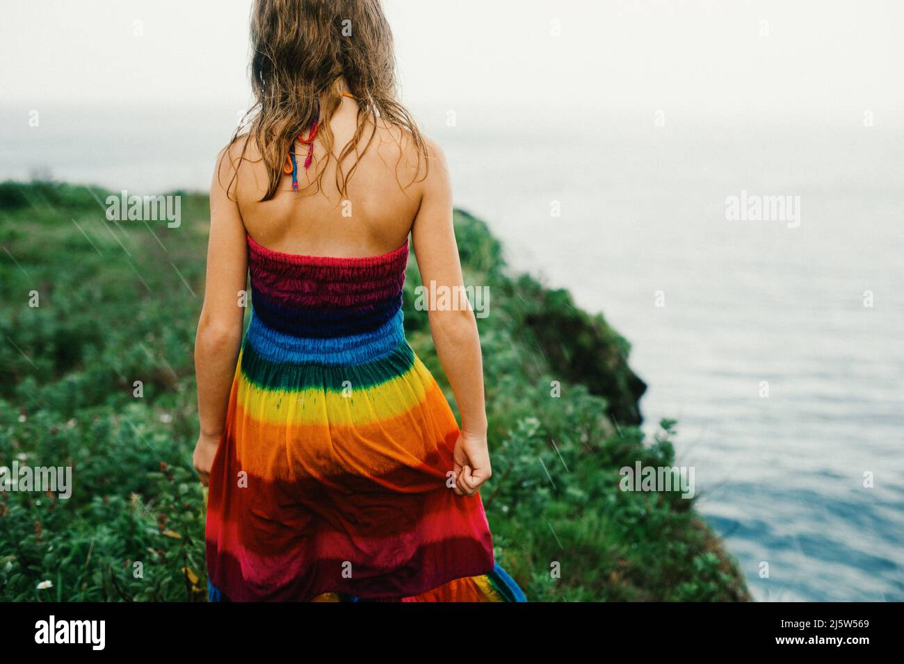 Mädchen läuft im Regen auf grünen Klippen am Meer in einem Regenbogenkleid Stockfoto