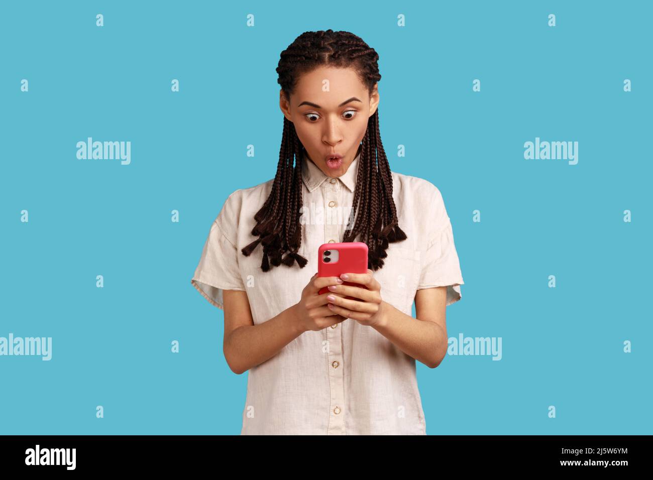 Emotional überraschte Frau mit schwarzen Dreadlocks starrt auf den Smartphone-Bildschirm, liest erstaunliche Nachrichten mit stauntem Gesicht, von etwas fassungslos. Innenaufnahme des Studios isoliert auf blauem Hintergrund. Stockfoto