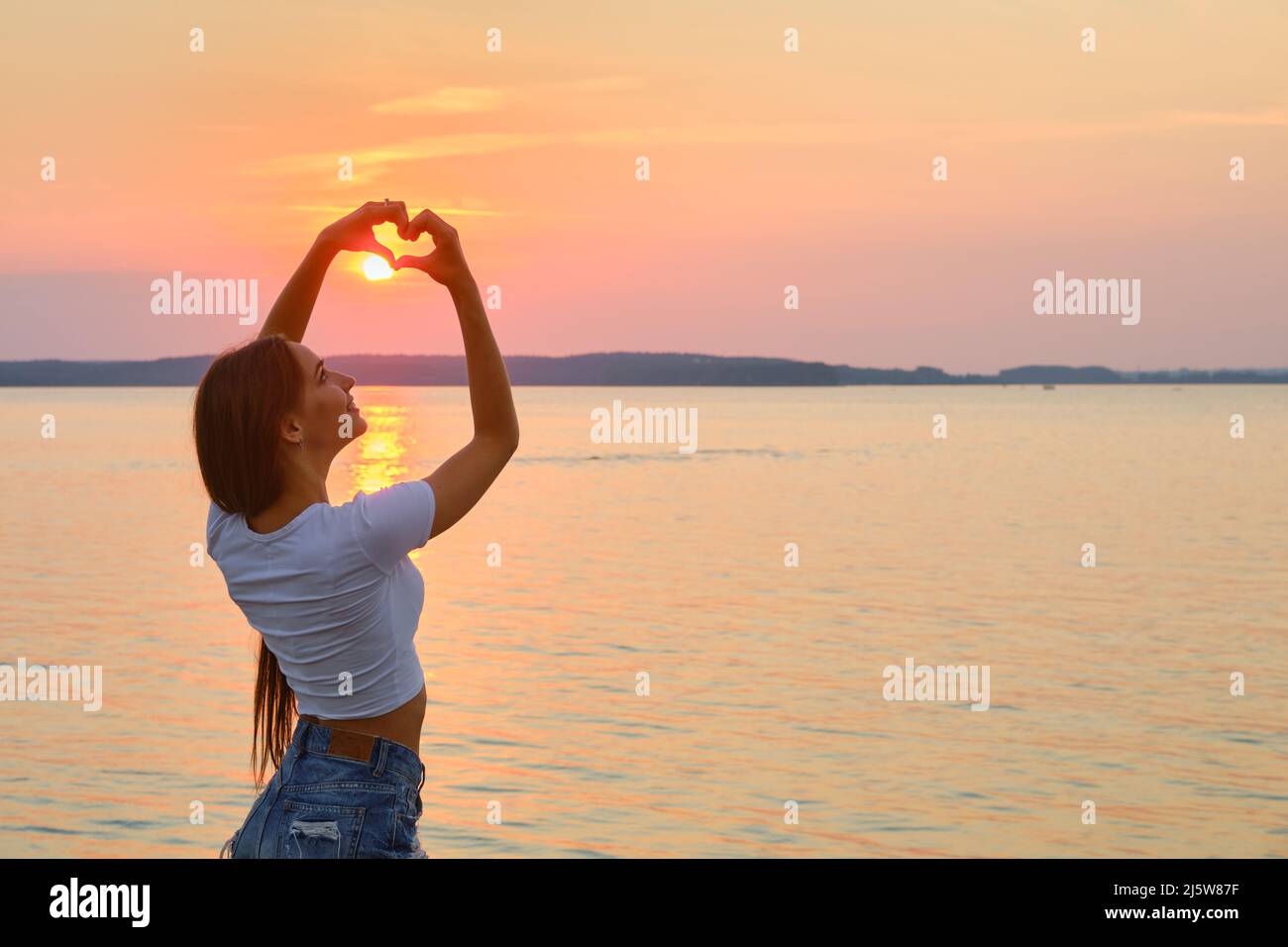 Die junge Frau zeigt mit ihren Armen ein Liebeszeichen. Sie legte die Finger in Herzform in Richtung Sonnenuntergang. Stockfoto