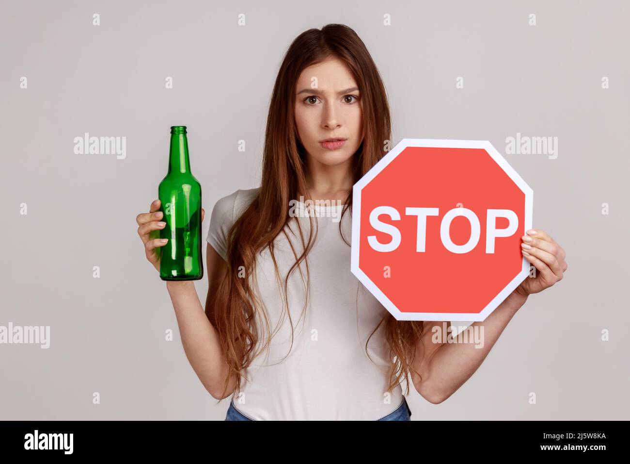Porträt einer ängstlich schönen Frau mit Bierflasche für alkoholische Getränke und Stoppschild, Warnung und Besorgnis, mit weißem T-Shirt. Innenaufnahme des Studios isoliert auf grauem Hintergrund. Stockfoto