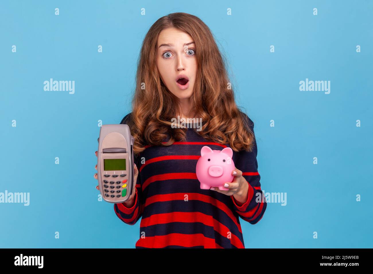 Die staunende Frau, die einen gestreiften Pullover im lässigen Stil trägt, POS-Zahlungsterminal und Smartphone mit leerem Bildschirm zeigt, hat den Ausdruck überrascht. Innenaufnahme des Studios isoliert auf blauem Hintergrund. Stockfoto