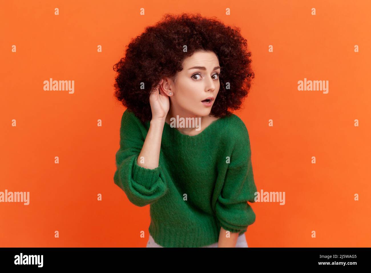 Frau mit Afro-Frisur trägt einen grünen Pullover im lässigen Stil, die Hand in der Nähe des Ohrs hält, um besser zuzuhören, Hörprobleme hat, schwer zu verstehen. Innenaufnahme des Studios isoliert auf orangefarbenem Hintergrund. Stockfoto