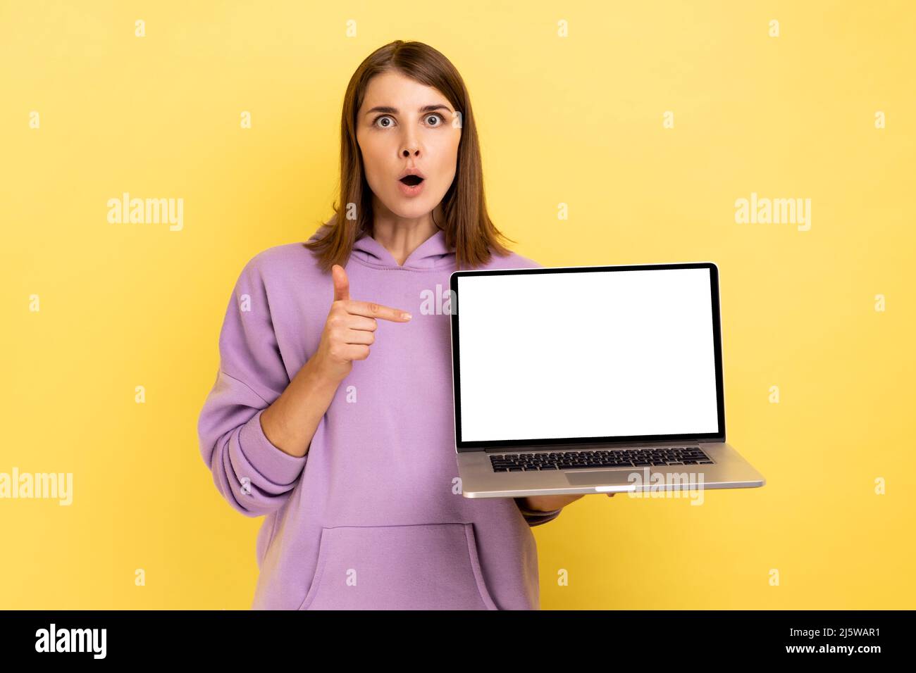Porträt einer überraschten dunkelhaarigen Frau mit großen Augen, die mit dem Finger nach oben zeigt und einen Laptop mit leerem Bildschirm hält, eine Idee hat und einen violetten Hoodie trägt. Innenaufnahme des Studios isoliert auf gelbem Hintergrund. Stockfoto