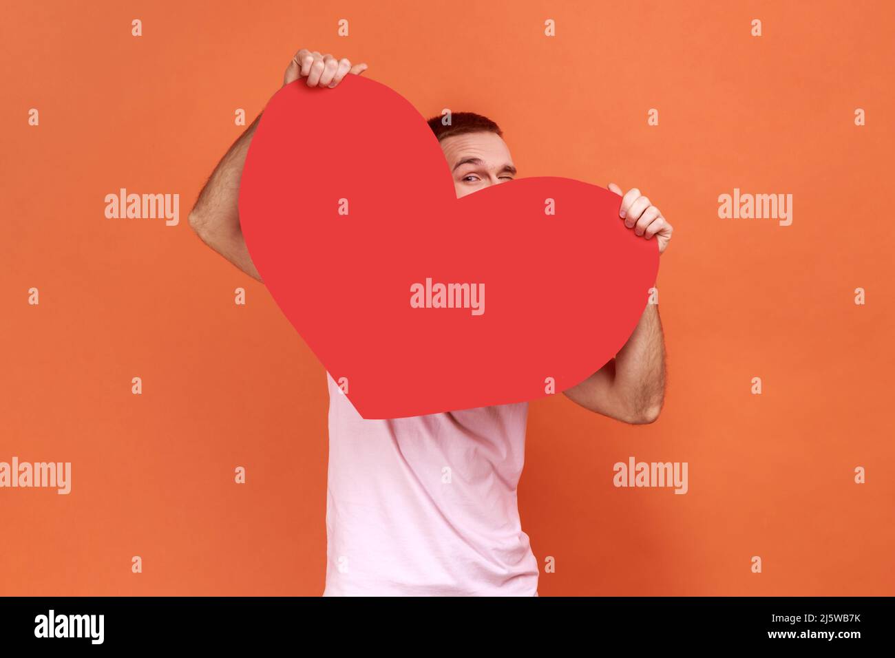 Porträt eines Mannes, der aus dem großen roten Herzen guckt und mit neugierigen neugierigen neugierigen Augen auf die Kamera blickt, Zuneigung und Zuneigung in einem pinken T-Shirt empfindet. Innenaufnahme des Studios isoliert auf orangefarbenem Hintergrund. Stockfoto