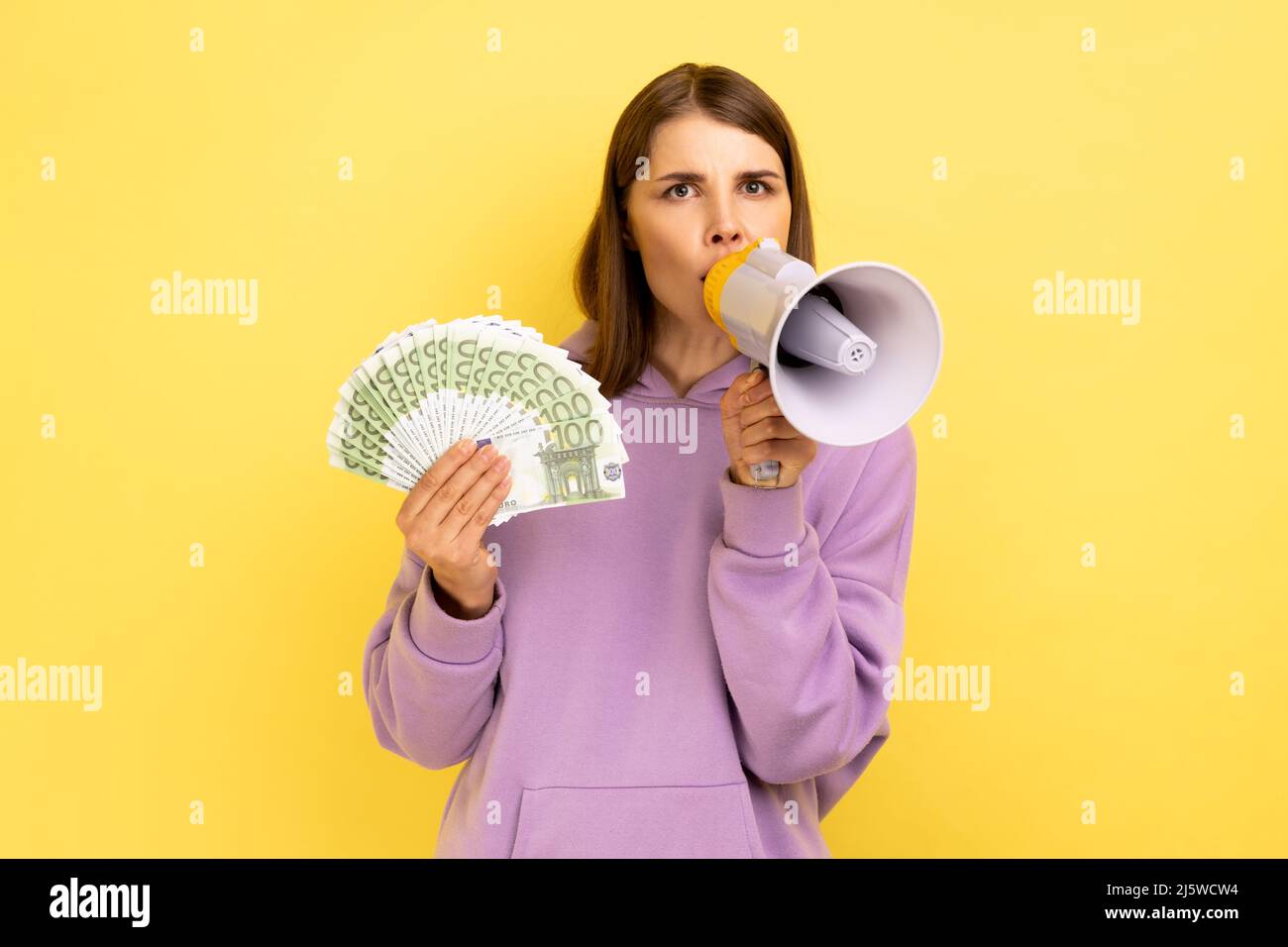 Porträt einer jungen erwachsenen Frau, die im Megaphon schreit und einen Euro-Fan hält, Boni und Aktionen ankündigt und einen violetten Hoodie trägt. Innenaufnahme des Studios isoliert auf gelbem Hintergrund. Stockfoto