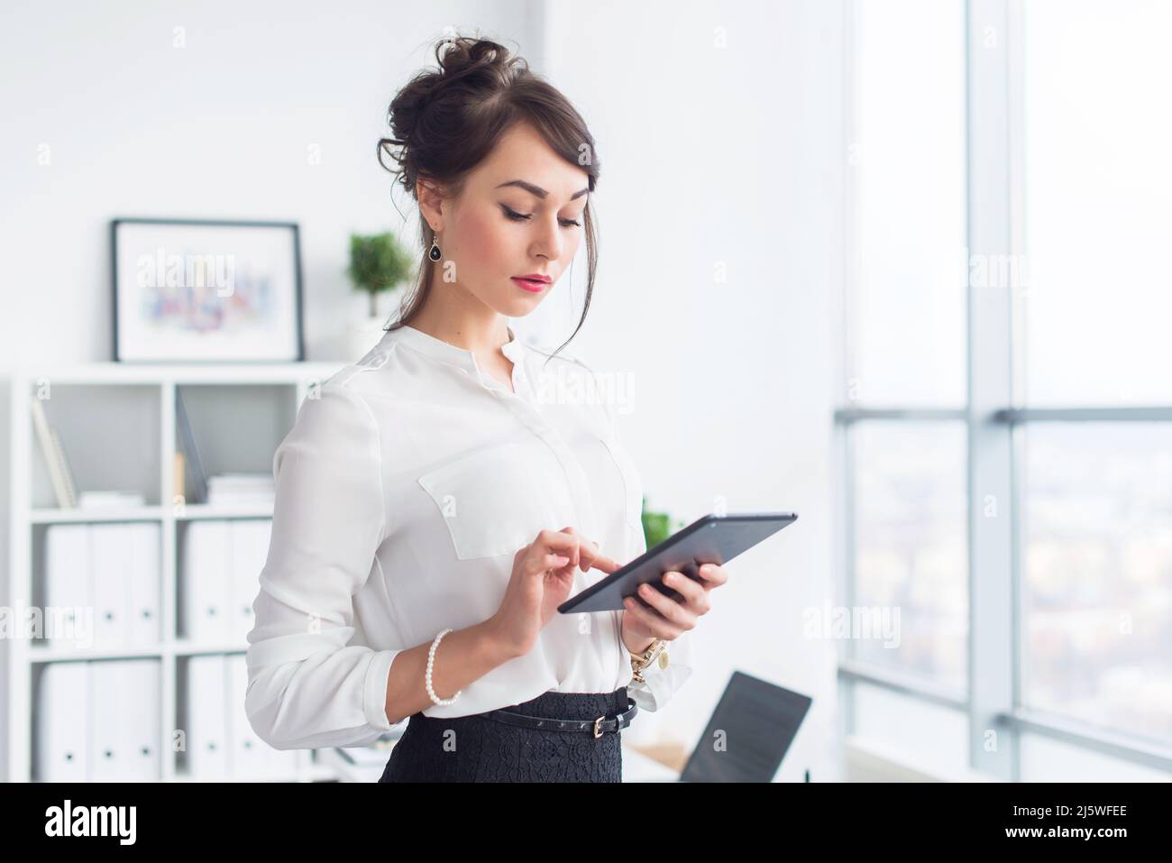 Junge lächelnde Büroangestellte an ihrem Arbeitsplatz liest und durchstöbert Nachrichten mit einem Tablet-Computer, während sie eine Pause macht Stockfoto