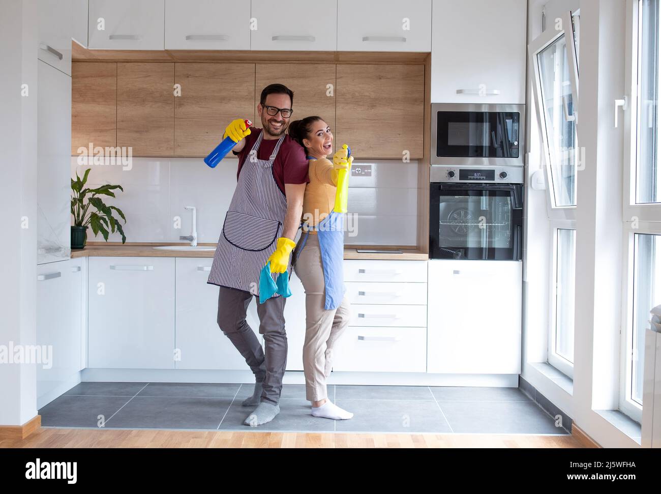 Junges Paar in der Küche mit Reinigungsmitteln, die Handschuhe tragen und ihre Aufgaben gemeinsam erledigen. Stockfoto