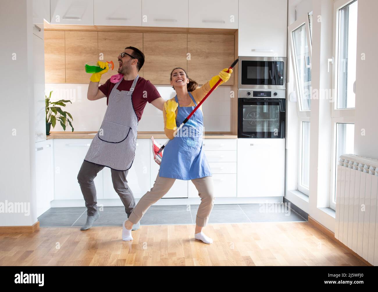 Fröhliches Paar tanzt und singt, während sie gemeinsam mit Reinigungsmitteln arbeiten, während sie Rockstars mit Sprühflasche und Mopp imitieren. Stockfoto