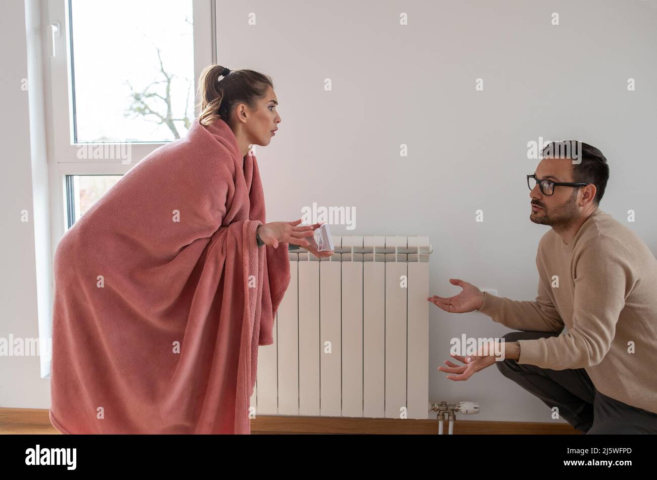 Frau mit einer Decke in der Wohnung bedeckt, die einem Mann eine niedrige Temperatur auf dem Thermostat zeigt. Stockfoto