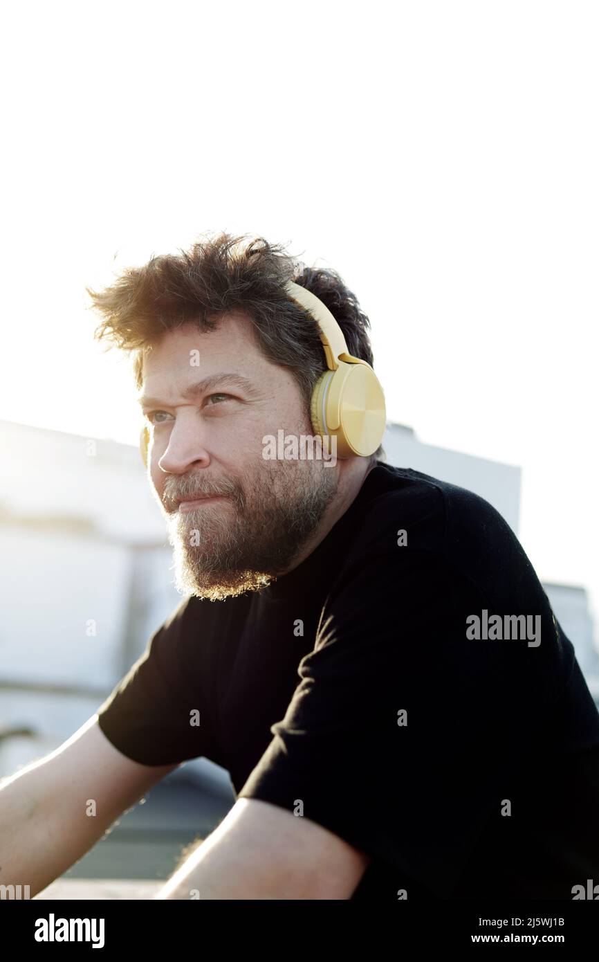 40-jähriger nordischer Mann mit Bart und Kopfhörern, der im Freien zur Seite blickt und das Abendlicht von hinten hat Stockfoto