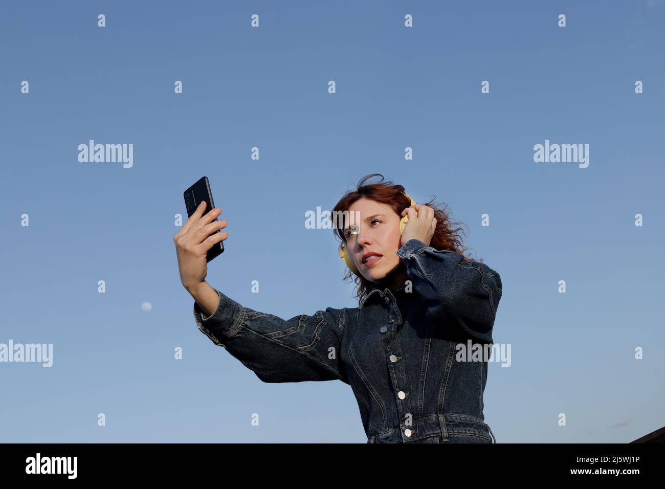 Kaukasische Frau mit Kopfhörern von unten, die auf ihren Handybildschirm mit einem tiefblauen Himmel blicken Stockfoto