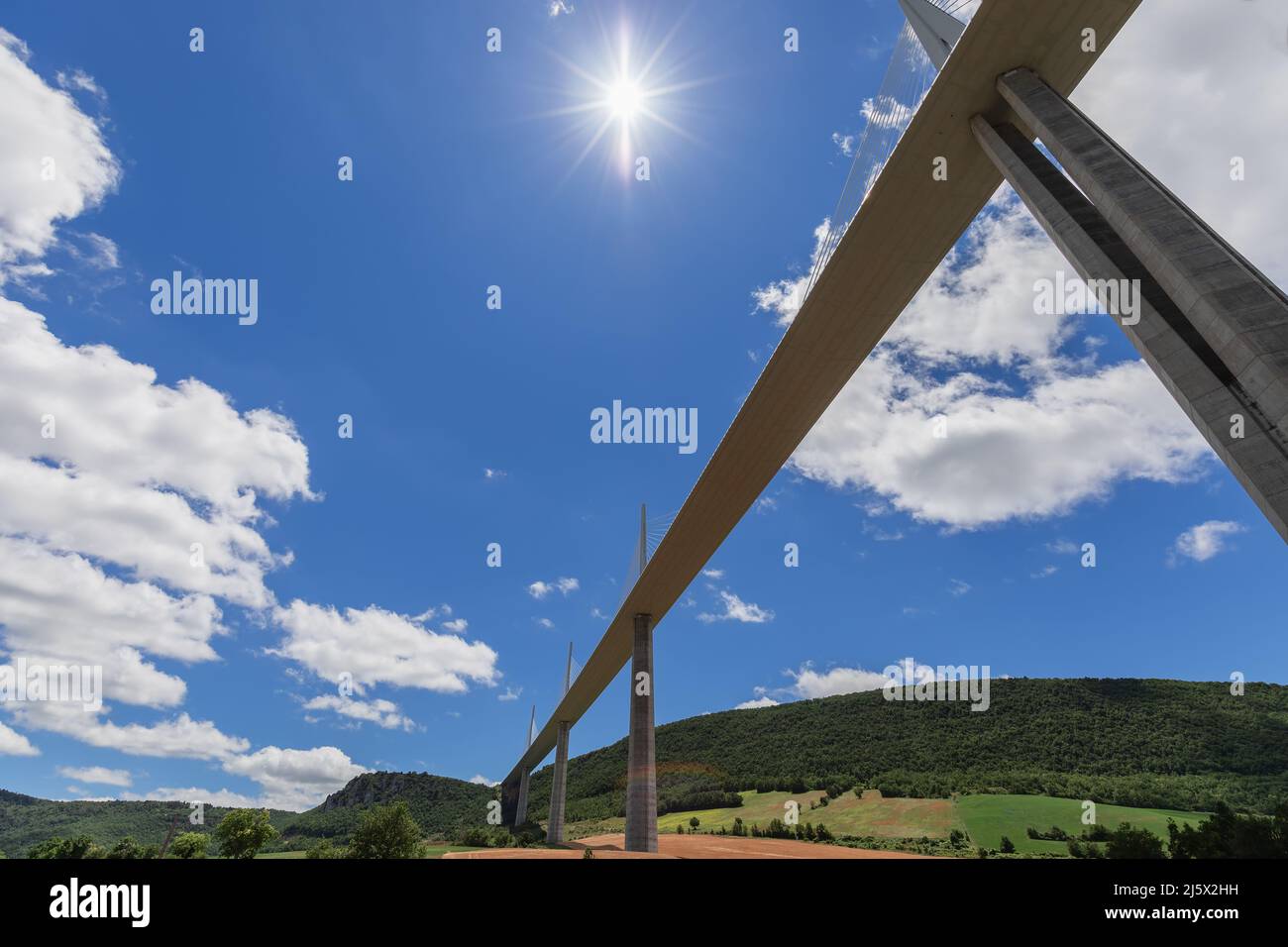 Die hellste Mittagssonne erhellt den blauen Himmel und das Viadukt von Millau, nahe gelegene landwirtschaftliche Felder, Wald auf Hügeln. Aveyron, Ozitanien, Südfrankreich Stockfoto