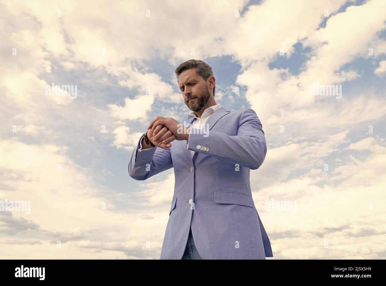 Schöner reifer Mann Geschäftsmann in Anzug Check-Zeit auf Armbanduhr auf Himmel Hintergrund, Pünktlichkeit. Stockfoto