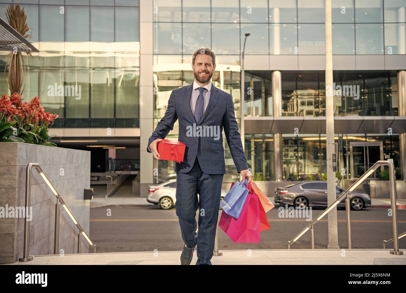 Glücklicher Unternehmer in Geschäftsanzug mit Einkaufstaschen Geschenke gehen vor dem Büro, Herrentag Stockfoto
