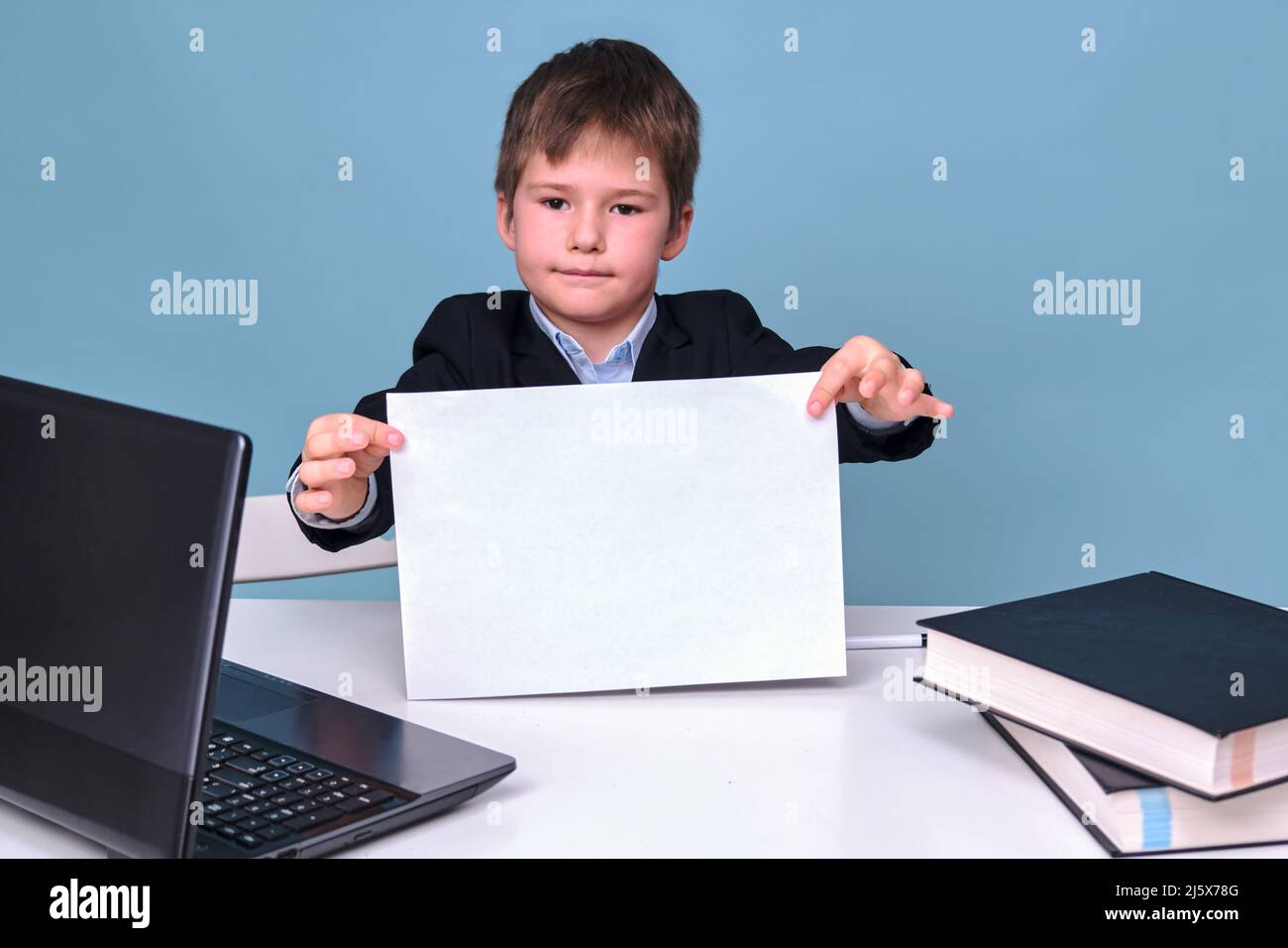 Ein Junge in einem Schulanzug mit einem Blatt A4 Papier an einem Computer während des Fernlernens, kopieren Sie Platz auf einem blauen Studiohintergrund Stockfoto