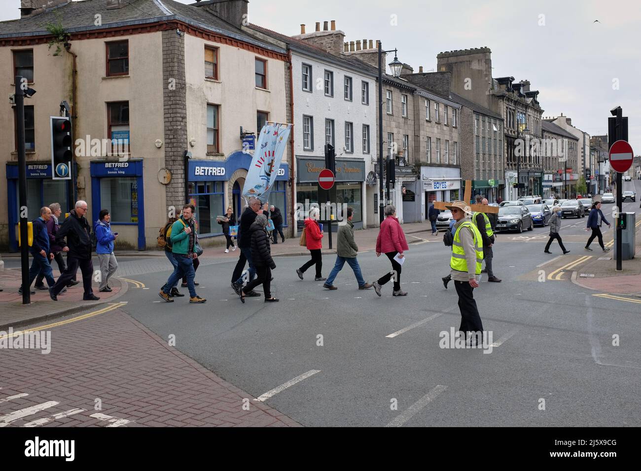 Osterprozession mit einem Transparent, das an der Ampel in Kendal, Cumbria, Großbritannien, die Straße überquert, wobei ein Mann in einer Hi-viz-Jacke anschaut Stockfoto