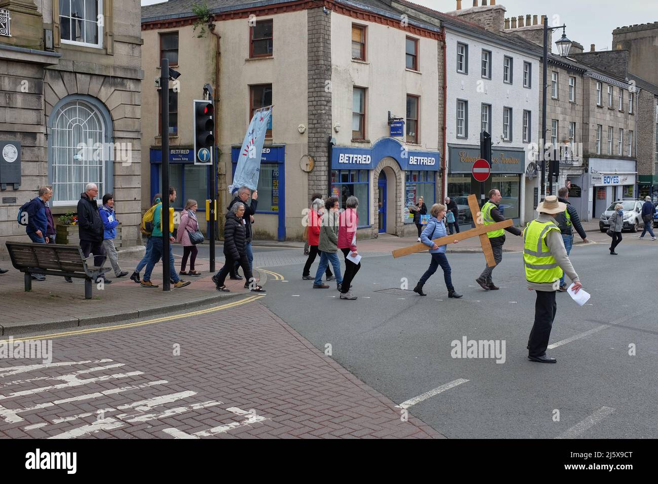 Osterprozession mit einem großen Kreuz und einem Banner, das die Straße an der Ampel in Kendal, Cumbria, Großbritannien, überquert, wobei ein Mann in einer Hi-viz-Jacke anschaut Stockfoto