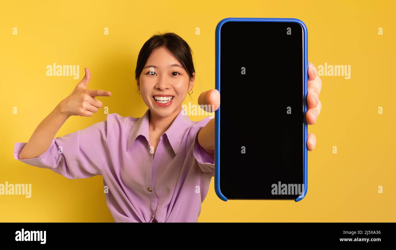 Tolle mobile App. Junge asiatische Frau zeigt auf Mobiltelefon mit leerem Bildschirm, Werbe-Website, Mockup Stockfoto