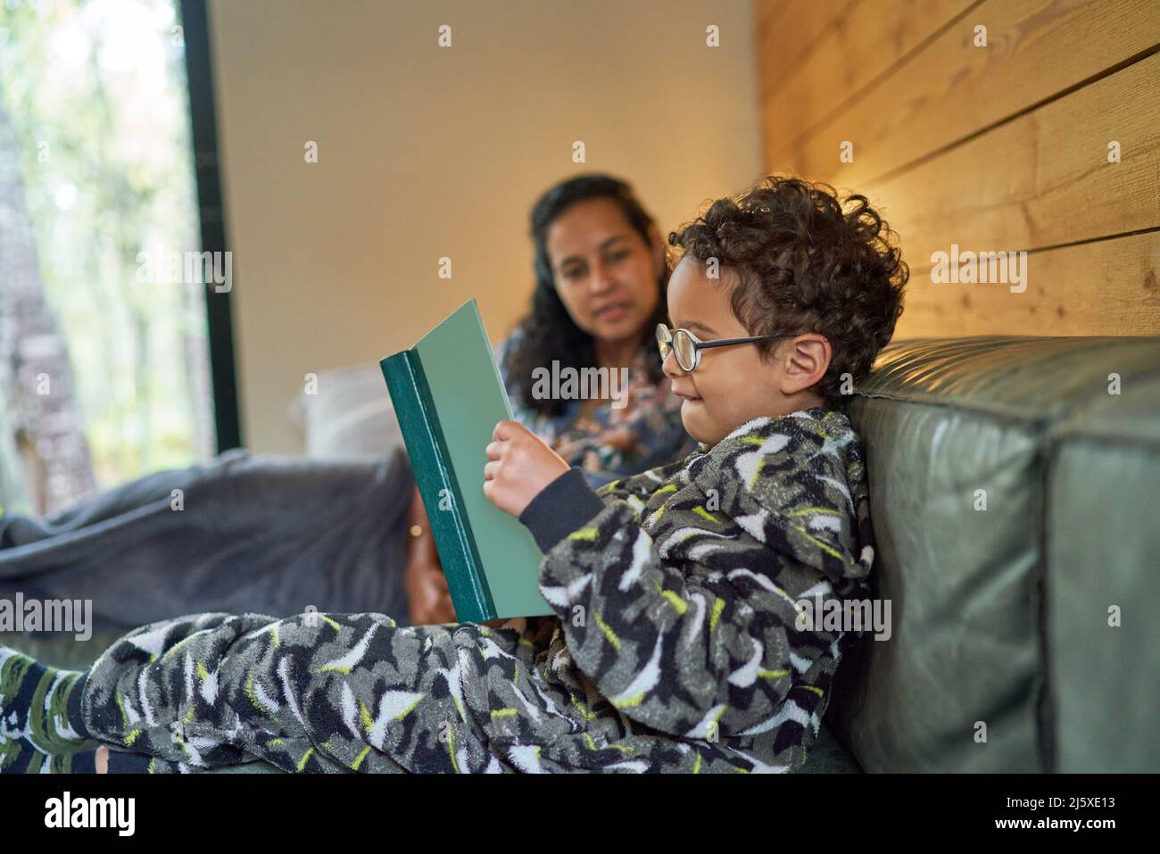 Mutter beobachtete den Sohn im Schlafanzug beim Lesen des Buches auf dem Sofa Stockfoto