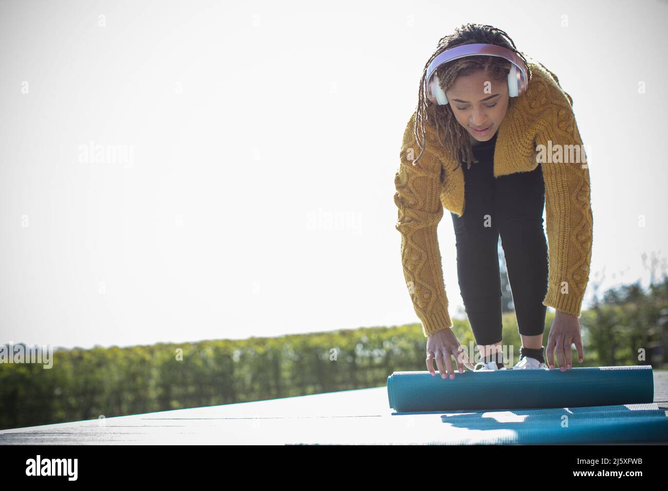 Junge Frau mit Kopfhörern, die auf der sonnigen Terrasse eine Yogamatte hochrollt Stockfoto