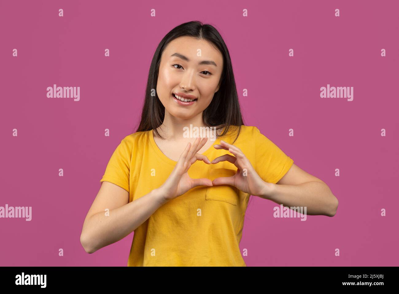 Glückliche asiatische Dame, die Herzgeste mit Händen in der Nähe der Brust zeigt, Liebeszeichen macht und auf violettem Studiohintergrund posiert Stockfoto