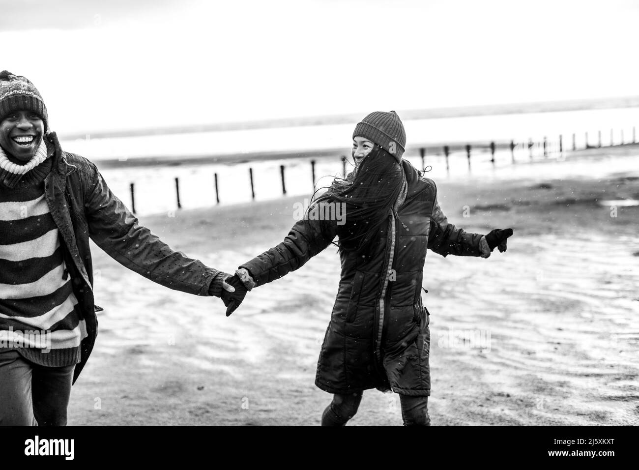 Verspieltes Paar in warmer Kleidung, das die Hände am Winterstrand hält Stockfoto
