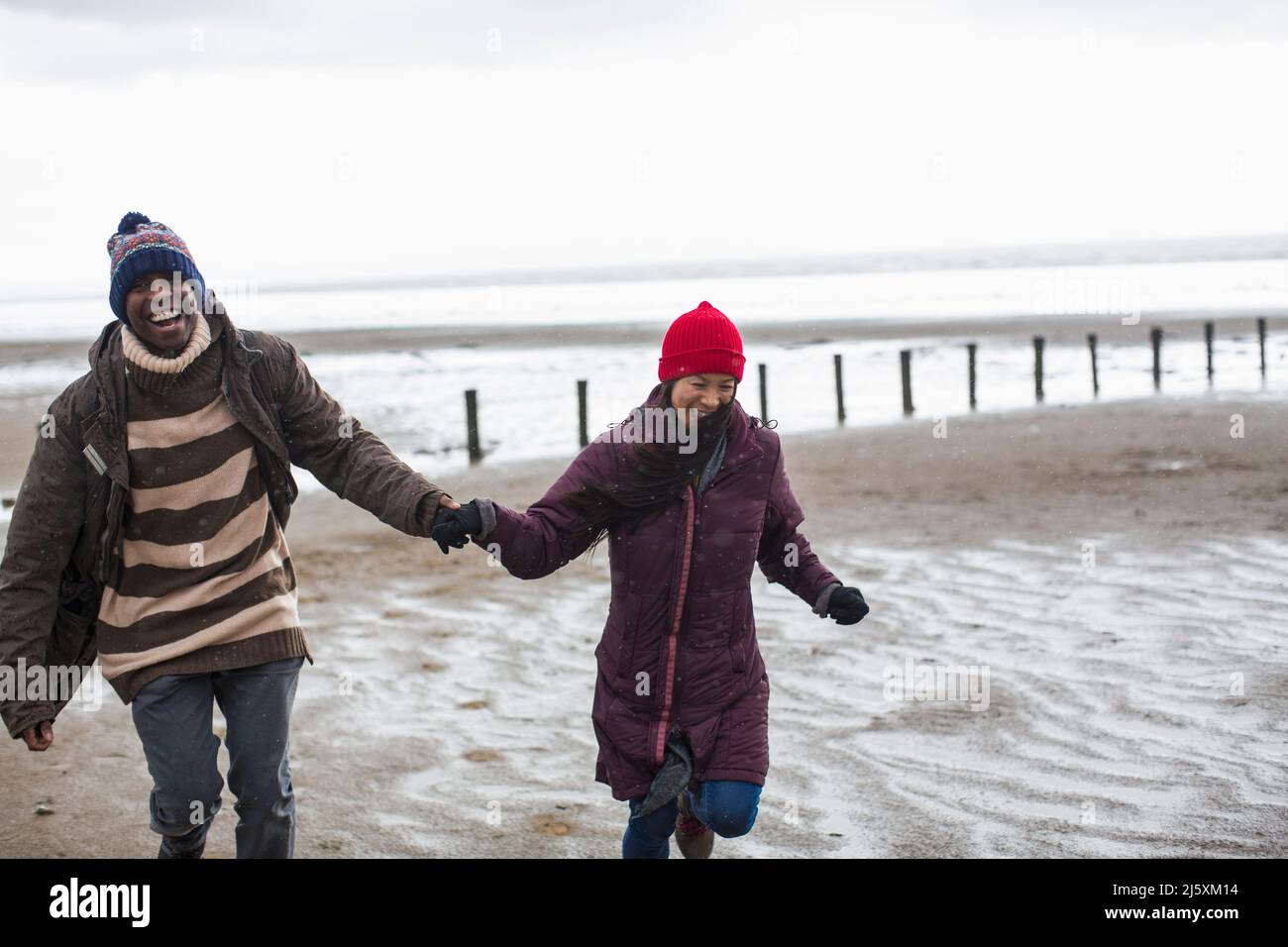 Glückliches sorgloses Paar, das die Hände hält und am nassen Winterstrand läuft Stockfoto