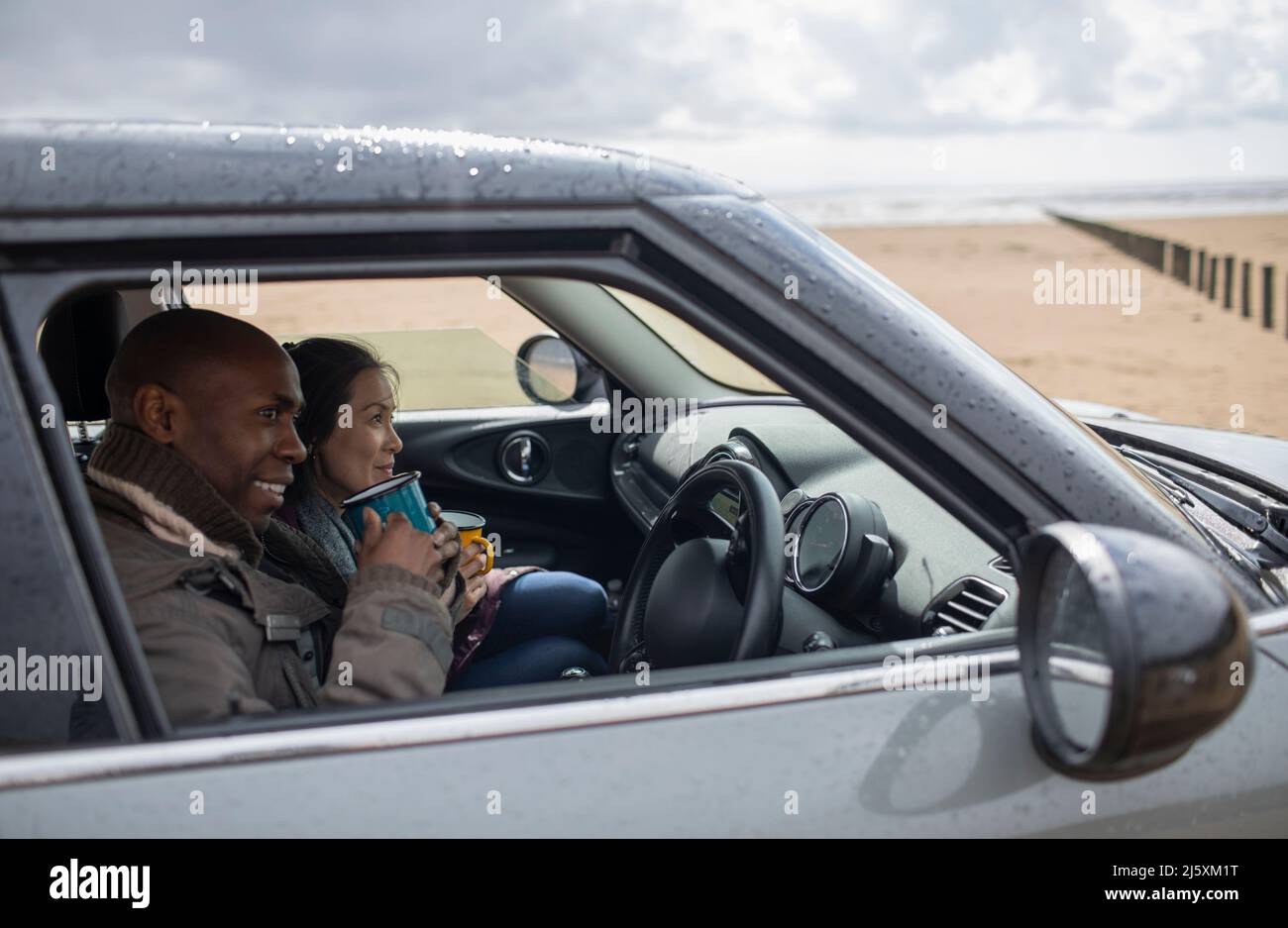 Glückliches Paar, das im Auto am nassen Winterstrand Tee trinkt Stockfoto