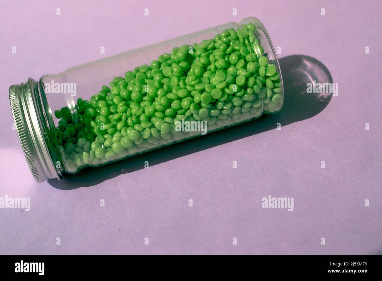 Grünes Wachs zur Enthaarung in Granulat. Auf violettem Hintergrund. Glas mit Wachs auf isoliertem Hintergrund. Stockfoto