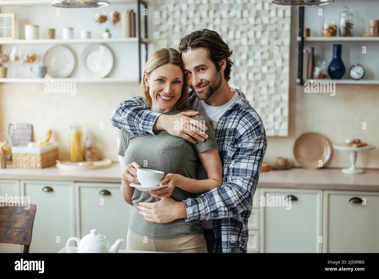 Lächelnder junger europäer mit Dreitagebart umarmt Dame von hinten mit einer Tasse Kaffee in der modernen Küche Stockfoto