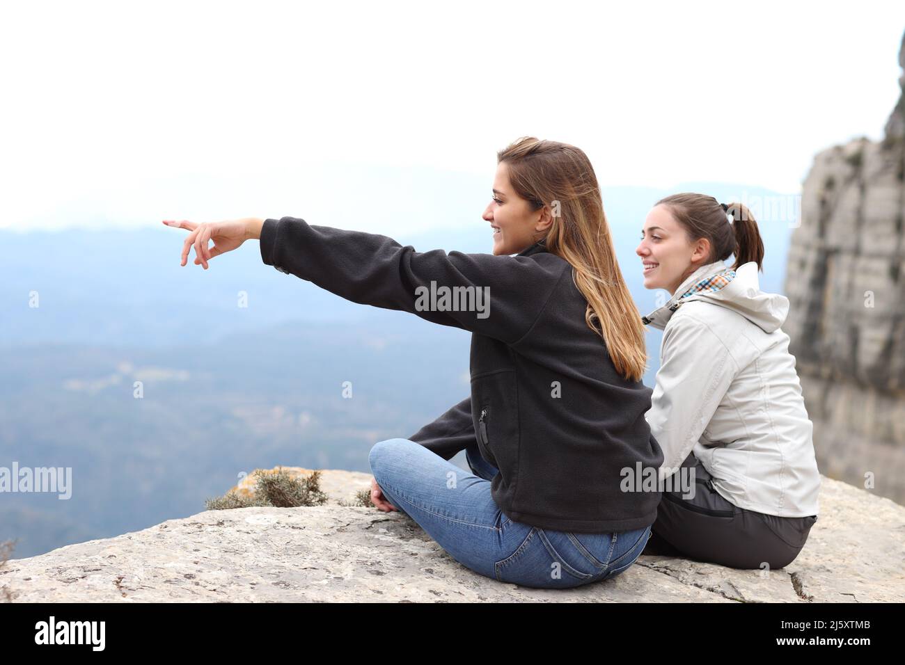 Zwei glückliche Wanderer, die auf einer Klippe sitzen und die Aussicht auf den Berg im Freien genießen und wegzeigen Stockfoto