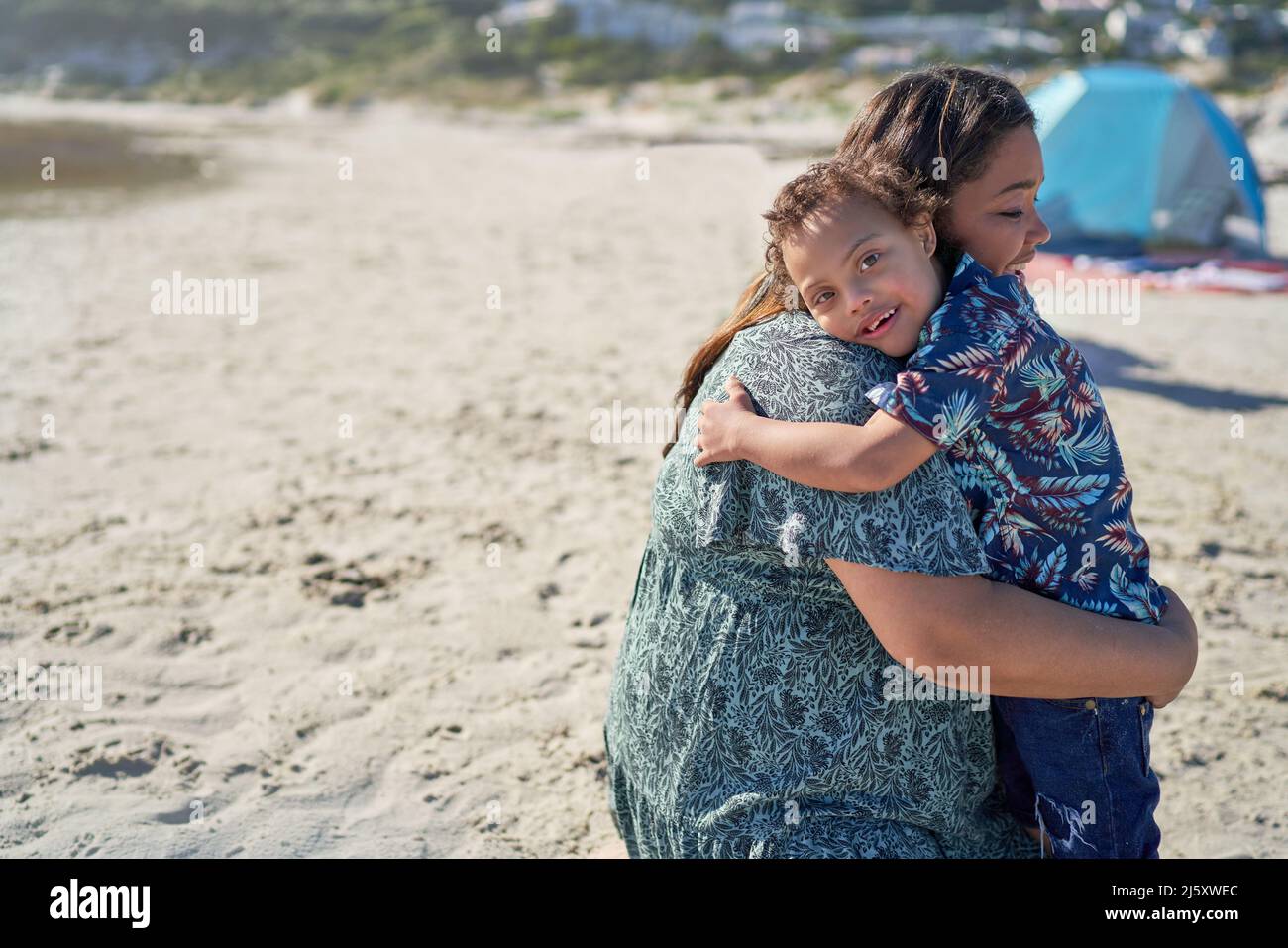 Portrait glücklicher Junge mit Down-Syndrom umarmt Mutter am Strand Stockfoto