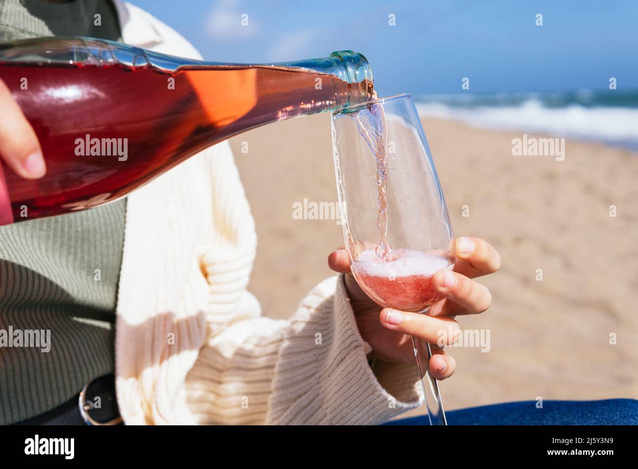 Erntet anonyme Frauen, die Rosenperlwein aus der Flasche ins Glas gießen, während sie am Sandstrand in der Nähe des winkenden Meeres im tropischen Resort sitzen Stockfoto